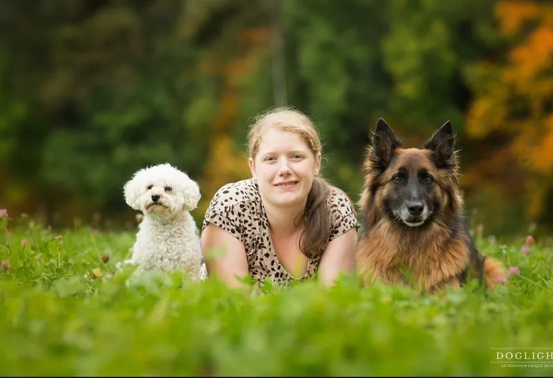 Tifanie, éducatrice canine CaniPlus, allongée dans l'herbe entre un bichon frisé et un berger belge tervueren